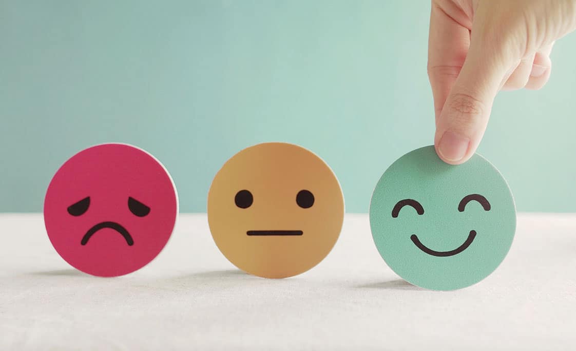 Foto de tres caritas: una triste, una seria y una feliz. Una mano escoge la feliz.