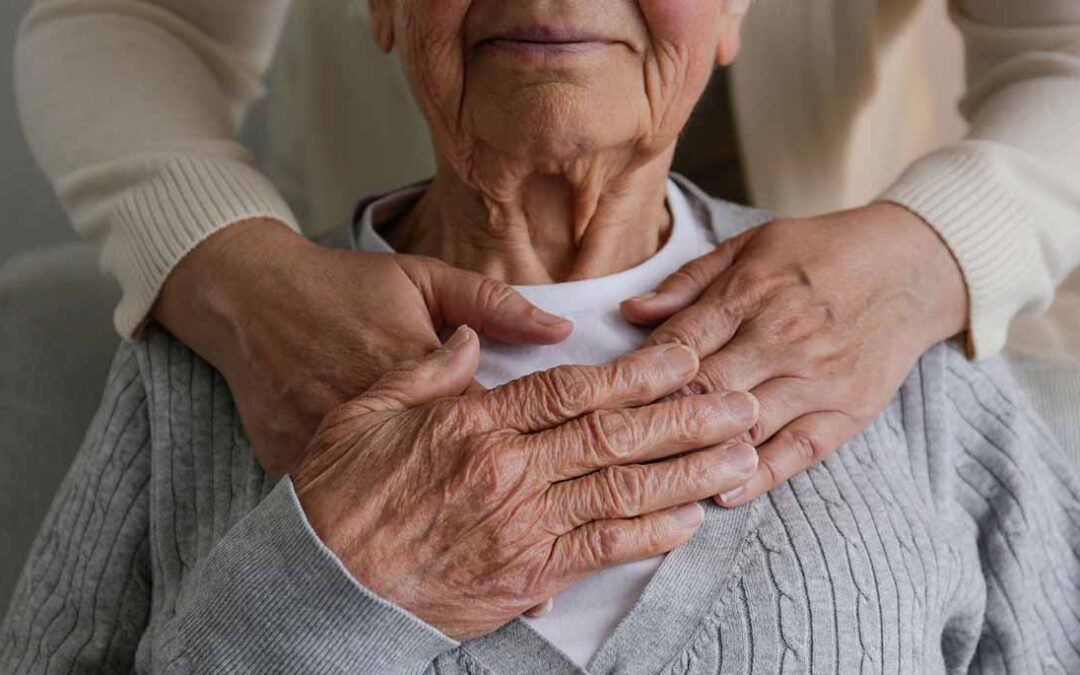 Una persona mayor se lleva la mano al pecho. Detrás una persona apoya sus manos en los hombros de la persona mayor.